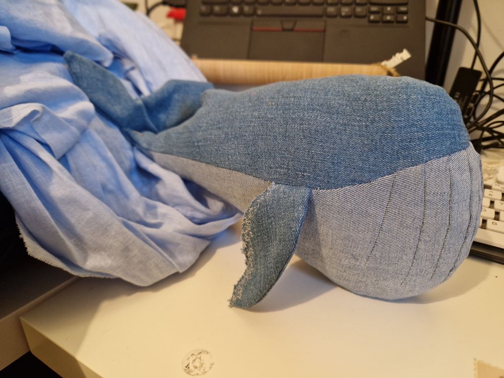 Toy whale with comically large ventral pleats (the chin folds on blue whales and their relatives) made from light blue denim on top and even paler blue on the belly. It doesn't have eyes yet and is balancing on a desktop with various things strewn around it