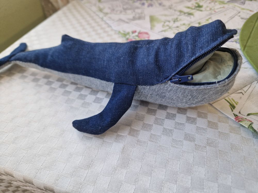 A maybe 20cm long toy whale made from denim fabric, deep blue op top and the pale inside at the bottom, gently stuffed. Its mouth is openable with a zipper, and reveals a pocket lined in pale green.