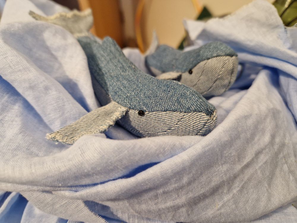 Two smaller toy whales, made from light blue top and even lighter bottom fabric, peeking out of a heap of different blue fabric with black bead eyes.