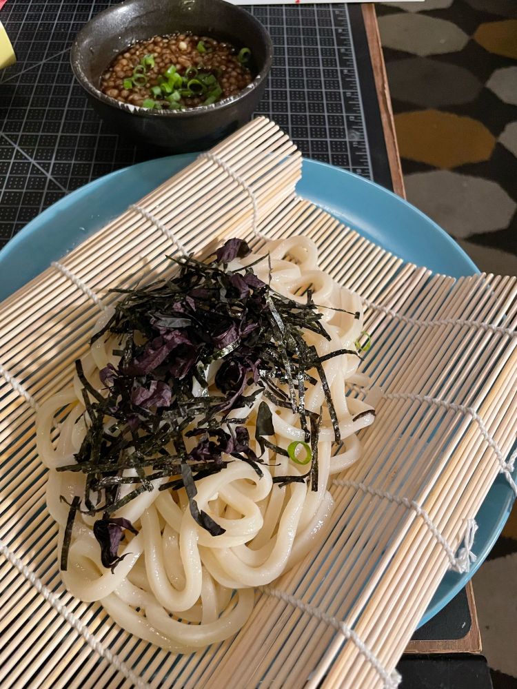 Pile of noodles with shredded nori seawees and shiso leaf, on top of a bamboo mat on a blue plate. Bowl of sauce sprinkled with green onion in the background.