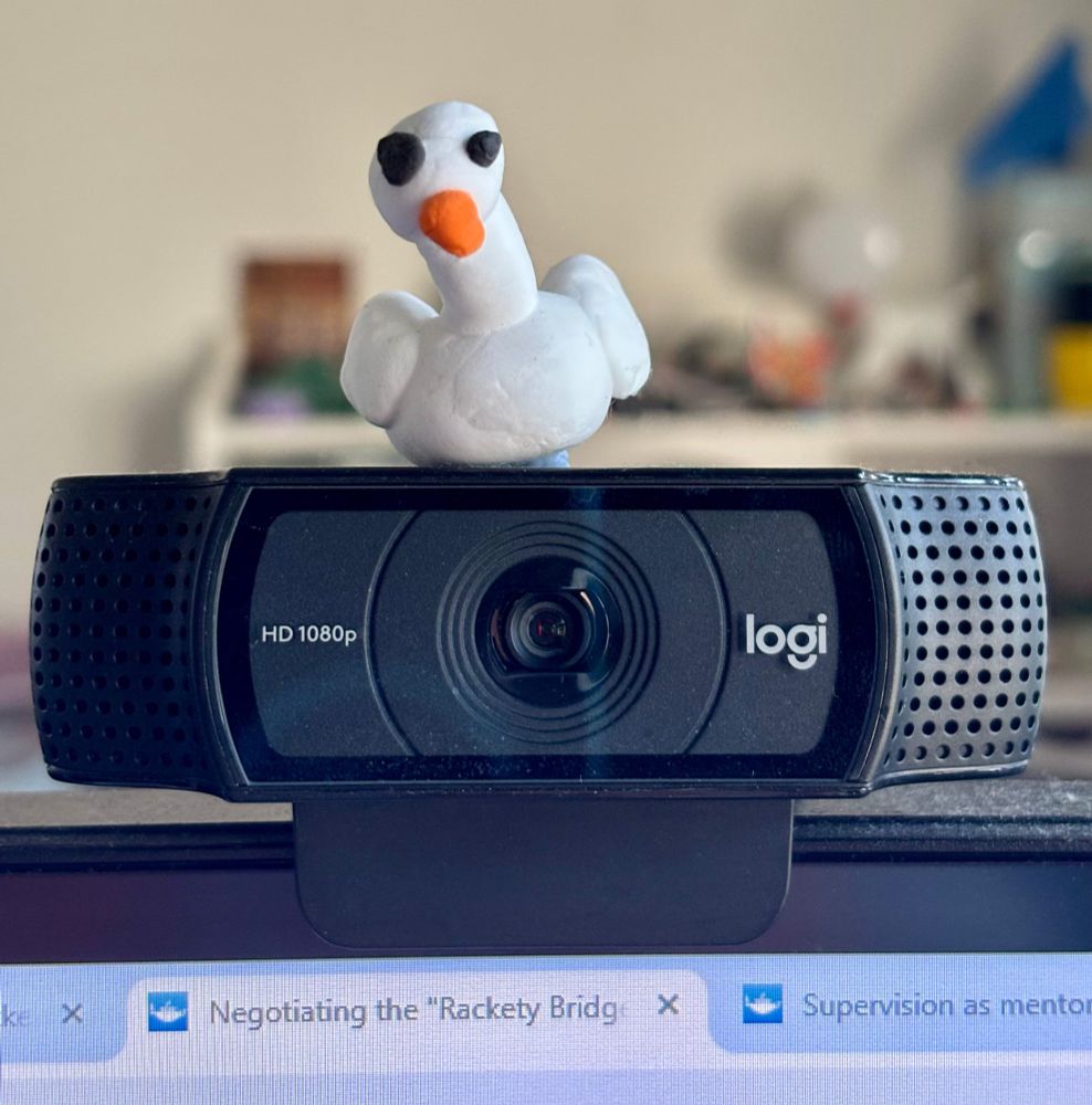 A tiny clay model of a goose sits upon a Logi brand webcam. It has a white body, black eyes, one bigger than the other, and an orange beak. The  neck is cocked slightly to its right, as if to convey attitude, or perhaps a lively cheerful manner. 