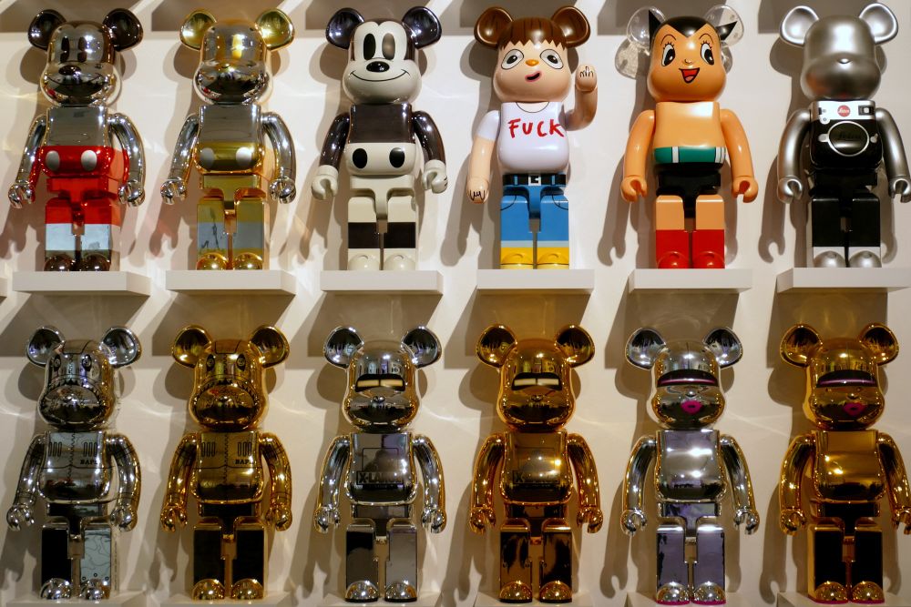 zweimal sechs ca. 50 cm hohe Kunststofffiguren, alle in der gleichen Grundform eines Bären mit Lego-Körper, stehen auf einzelnen Podesten nebeneinander vor einer weißen Wand. Der untere Teil ist wie goldene und silberne Roboter gestaltet, der obere Teil u.a. wie Micky Maus, eine Leica-Kamera oder ein Mädchen mit Stinkefinger und einem T-Shirt mit der Aufschrift FUCK. 