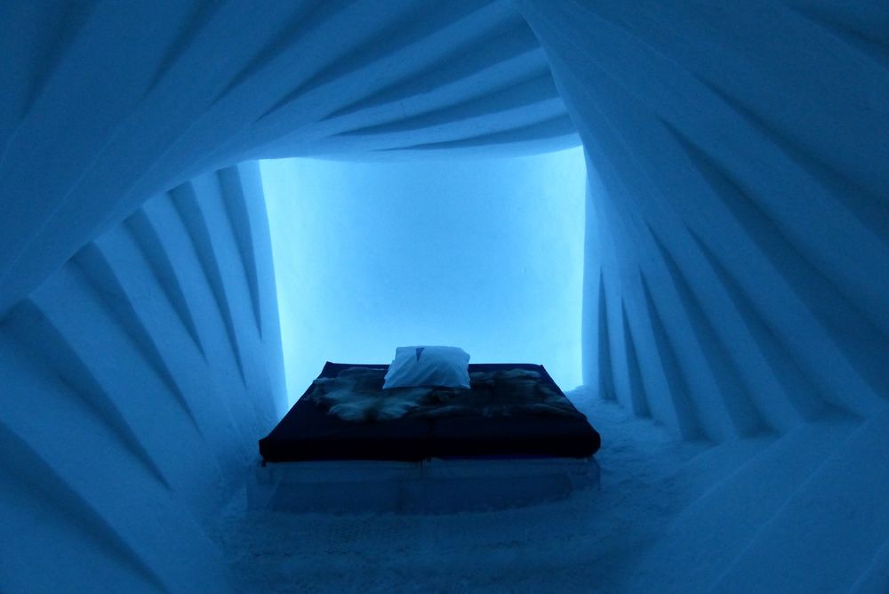 Zimmer in einem Eishotel. Dicke Felle auf einem Bett aus Eis. Der Raum besteht aus nach hinten kleiner werdenden Quadraten, die immer im leichten Winkel versetzt sind.
Das ganze Bild ist blau.