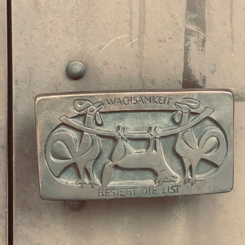 An einer Bronzetür befindet sich ein breiter Türgriff mit einem Bronzerelief: Zwei Hähne tragen eine Stange über der Schulter, an der ein Fuchs hängt. Titel: Wachsamkeit besiegt die List
