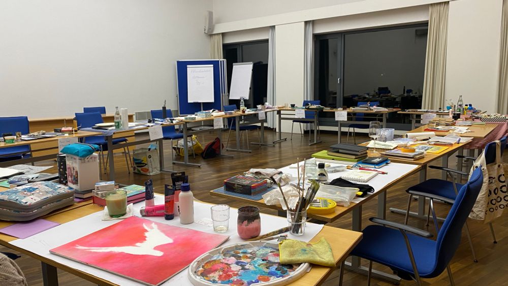 Ein weißer Raum mit Tischen im Quadrat  gestellt, aller voller Mal- und Zeichenkram