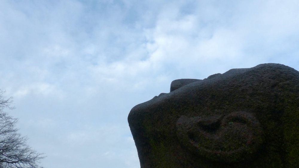 Blick von unten am Kopf einer Steinskulptur vorbei in den wolkigblauen Himmel. 