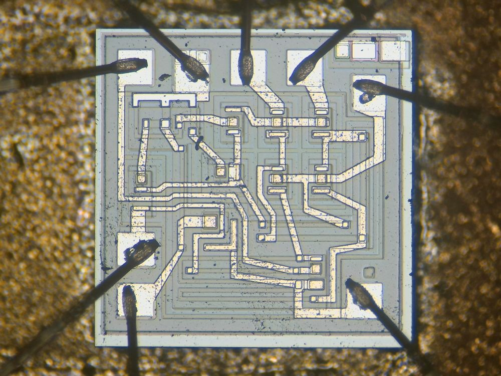 small rectangle of silicon against a gold background in soft focus. from the edges, eight bond wires shoot off in various directions. a delicate tracery of microscopic aluminum wire connects the nine transistors in the circuit. faint rectilinear outlines of diffusion regions can be seen -- these form transistors and resistors.