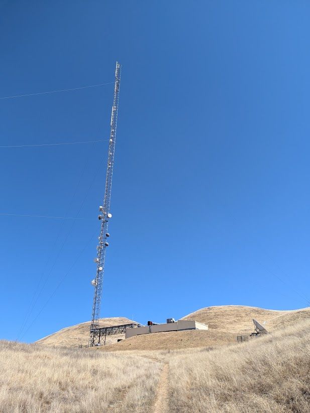 a large TV/radio antenna with multiple sets of antennas. clustered in the lower half are several microwave dishes. a low building houses the transmitters.
