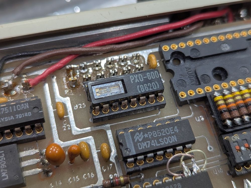 16-pin DIP in a circuit board. there's a square opening on the left with a glass-encapsulated tuning fork crystal. the date code is 8620, presumably meaning the 20th week of 1986.