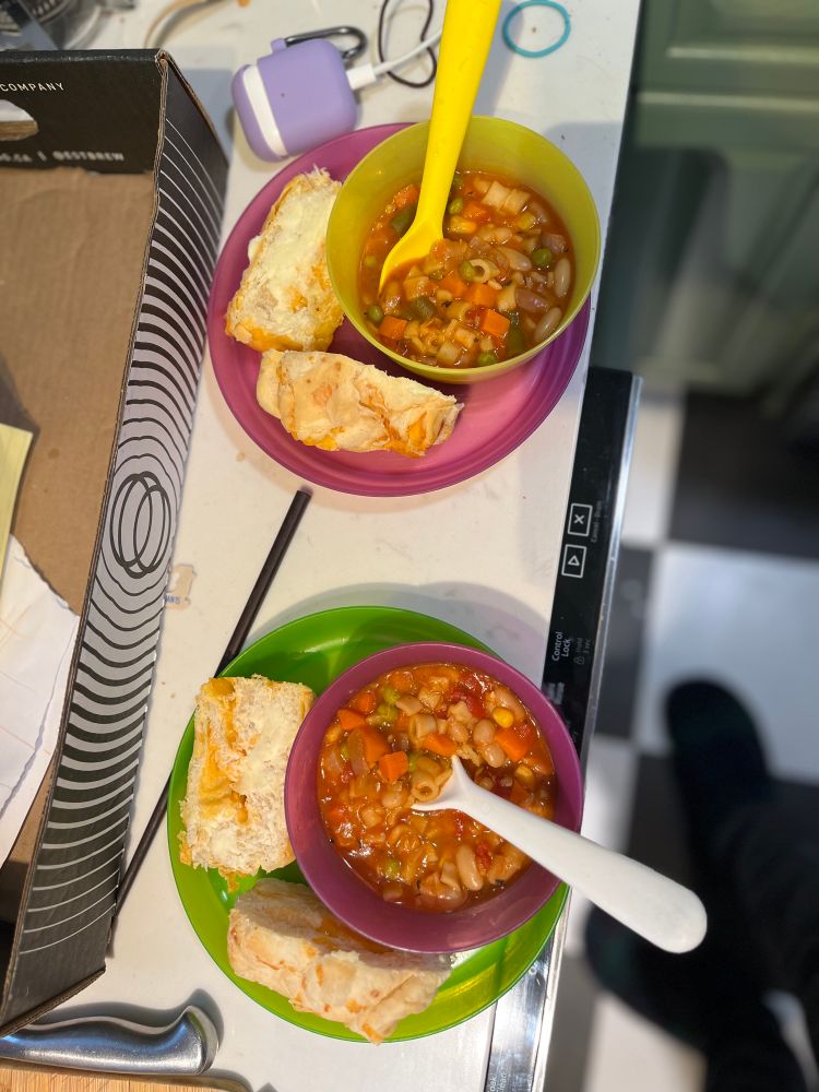 Two plastic bowls and plates featuring a meal of soup and cheese bun. 