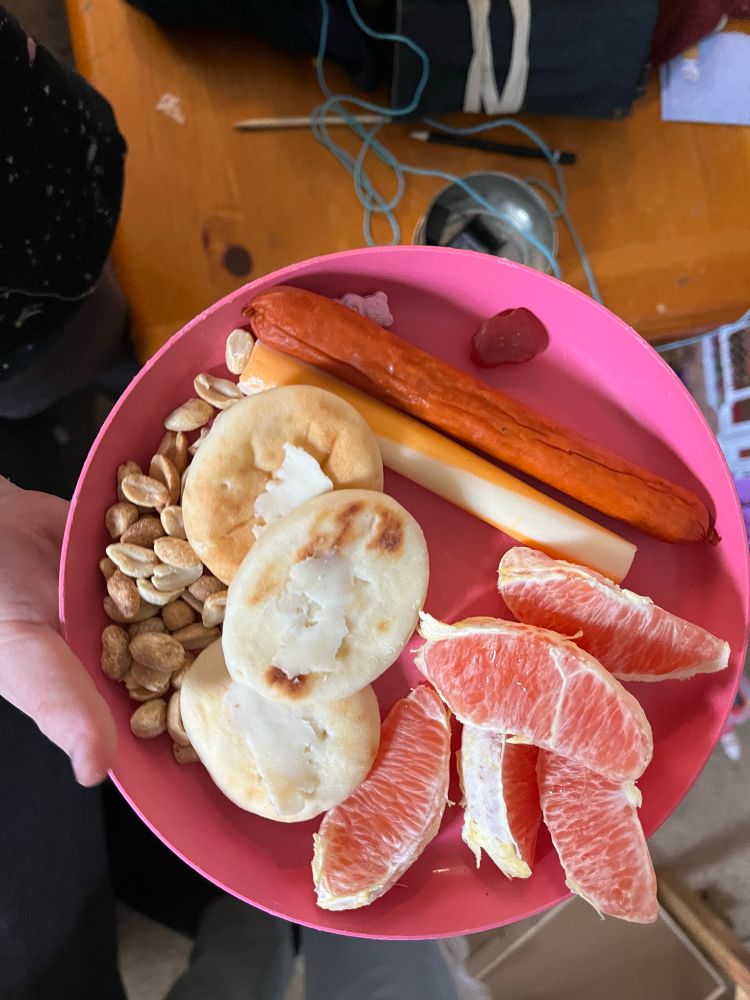 A meal of peanuts, mini pita bread, cheese string, pepperoni stick and orange segments on a pink plate. 