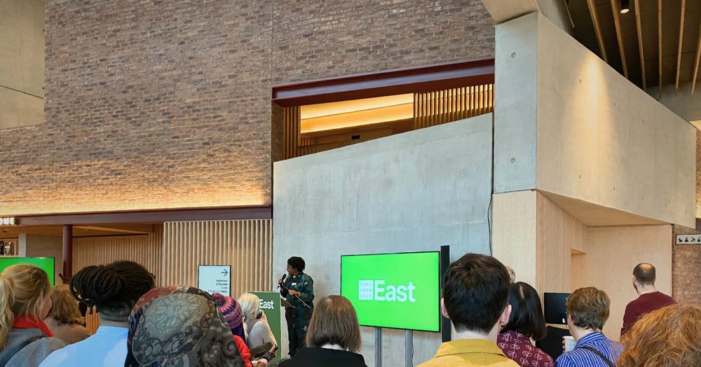 Natasha Anderson, head of Community Engagement at Sadlers Wells East, gives a speech in front of a diverse crowd in the newly opened venue. Next to her stands a screen displaying the Sadlers Wells East logo on a bright green background. 