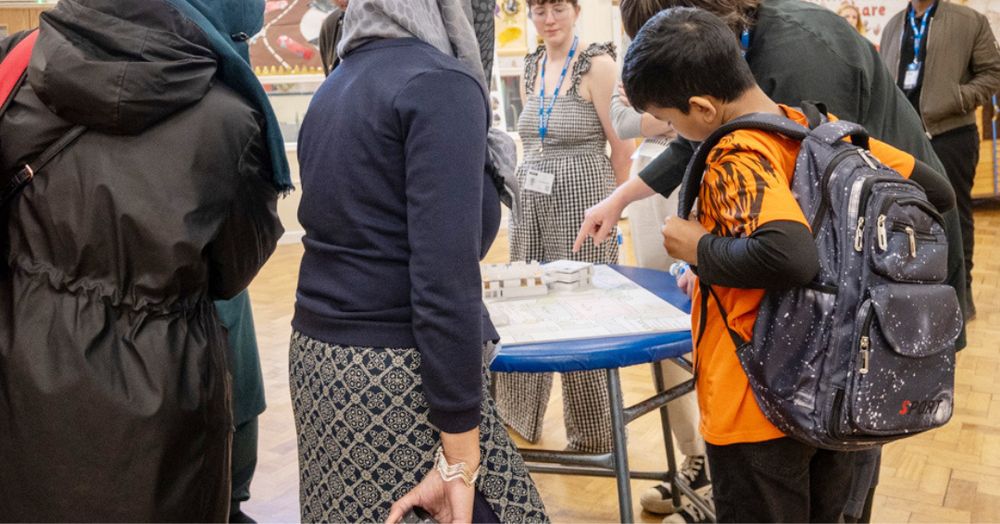 Staff, parents and a young child gather around a small architectural model of the new primary school, an member of the design team points at the roof of the school model, seemingly in the process of describing the plans to them. 