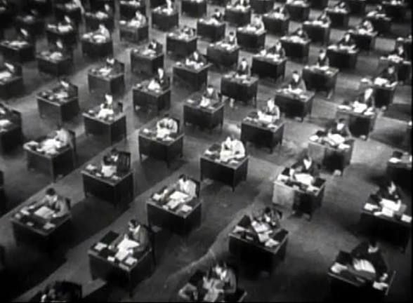 A black-and-white overhead shot of many people arranged symmetrically at many office desks. It’s a defining dystopian image from Orson Welles’s film “The Trial,” his 1962 adaptation of the 1925 Franz Kafka novel.