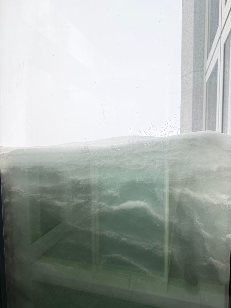 A shot from the inside of the Jungfraujoch building to the outside. The picture shows how the snow outside piles up against the window. On the right side, a small part of the outside front is visible. Since visibility is only a few meters due to bad weather conditions, there is no sky but there's only white space from fog and snow.