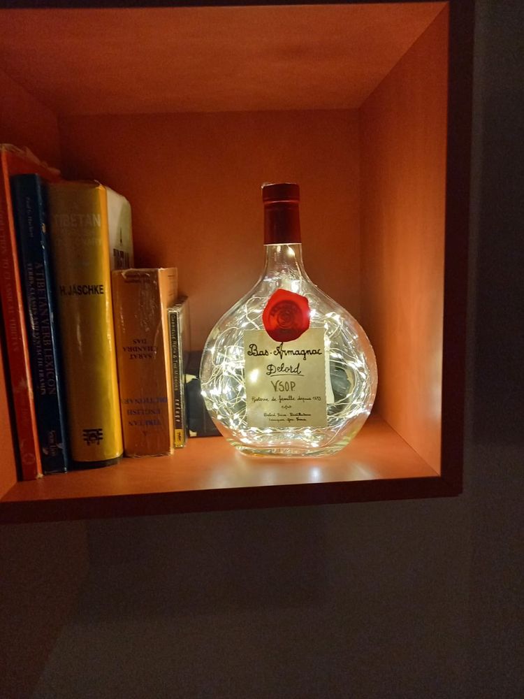 An empty Armagnac bottle with fairy lights inside, sitting on a shelf next to some books.