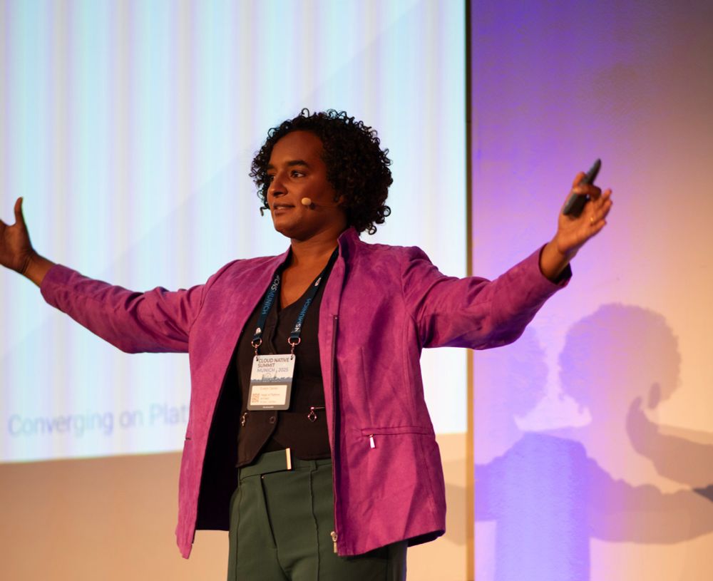 Standing on a stage with my arms out while speaking, I’m wearing a head piece and a clicker in one hand. I have an oversized purple jacket on over a dark blue vest and green pants. My curly hair has been swept back from my gesturing and looks slightly disheveled 