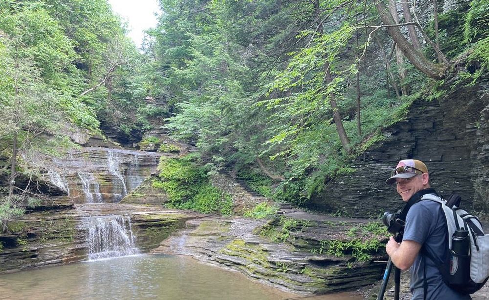 Me at one of the buttermilk falls cascades looking back towards Laura’s camera. I’m holding my large tripod and camera looking at a layered rock gorge with a 20’ wide pool at the base and forest greenery at the side and top of the gorge.
