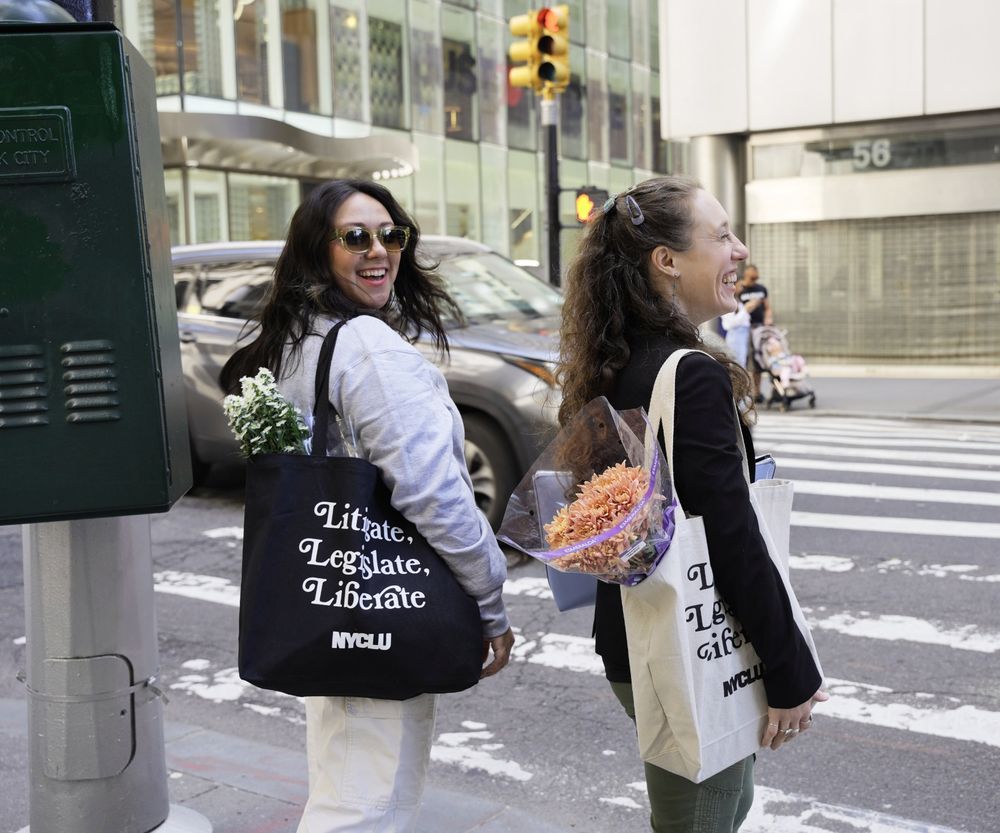 Two women stand at a crosswalk. One has dark hair and is wearing sunglasses. She is turned to face the camera and has a black NYCLU tote bag with flowers over her shoulder. The other woman has light brown hair with a barette and is laughing facing away. She has a canvas NYCLU tote over her shoulder with flowers inside.
