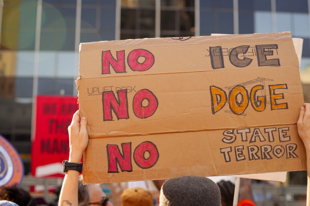 A person holds a handmade cardboard sign in the air at a protest. The sign reads "NO ICE, NO DOGE, NO STATE TERROR"