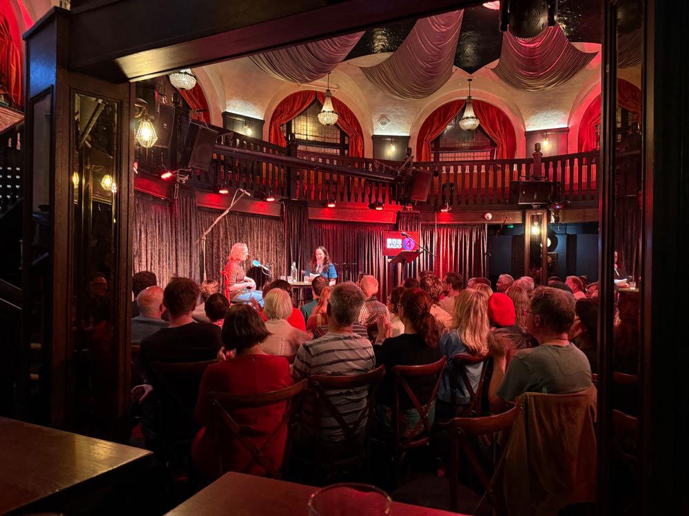 Two comedians on stage for a taping of a BBC radio 4 show. It’s taped in front of a live studio audience. 