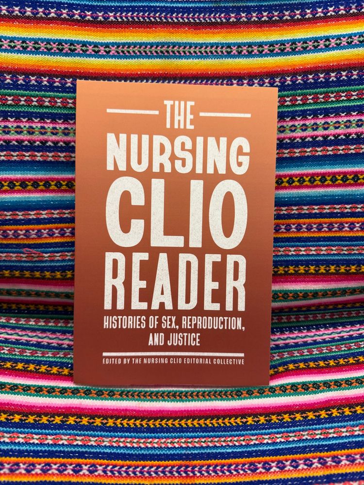 An orange paperback book titled “The Nursing Clio Reader: Histories of Sex, Reproduction, and Justice,” set against a background of colorful woven cloth from Peru. 