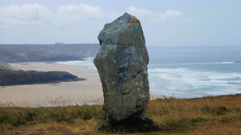 A large single standing stone, high on a cliff, overlooking a large sandy beach with braking Atlantic waves rolling in.