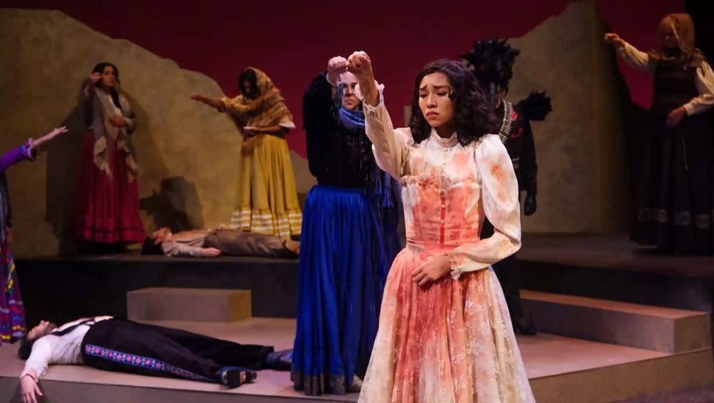 A photo from a production of Blood Wedding; every woman in the cast drops salt on the stage; the women wear long, colorful dresses in the style of the Mexican Revolution; the woman in the foreground wears white and is covered in stage blood. 