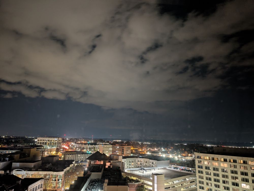 A long exposure photo of night in an American city. Clouds drift overhead, lit up from below by the multitude of lights on the ground. In the left of the photo cars' taillights blur together creating glowing arcs of light. 