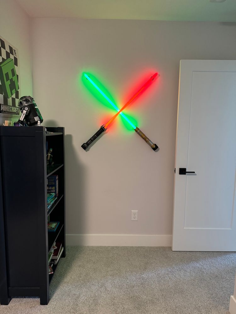 Two Savi’s Workshop lightsabers, one power and control saber in black with a red blade, and the other is peace and justice saber with a green blade. They are on a soft grey wall between a blue bookcase and a white door.