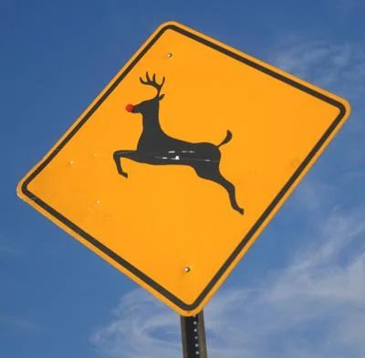A photo of a yellow street sign with the outline of a deer as a warning for deer crossings. The deer outline has a medium sized red sticker placed where the deer's nose would be. The sign has a blue sky behind it with some small whispy clouds in the lower right corner of the photo.