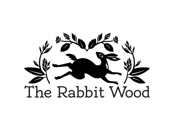 A black and white graphic logo featuring a rabbit in the center with legs outstretched as if running or hopping and it's head swiveled to look behind itself and up. The rabbit is wreathed in decorative leaves and flower buds in a semi-circle above and to it's sides. Underneath the decoration is the type "The Rabbit Wood"