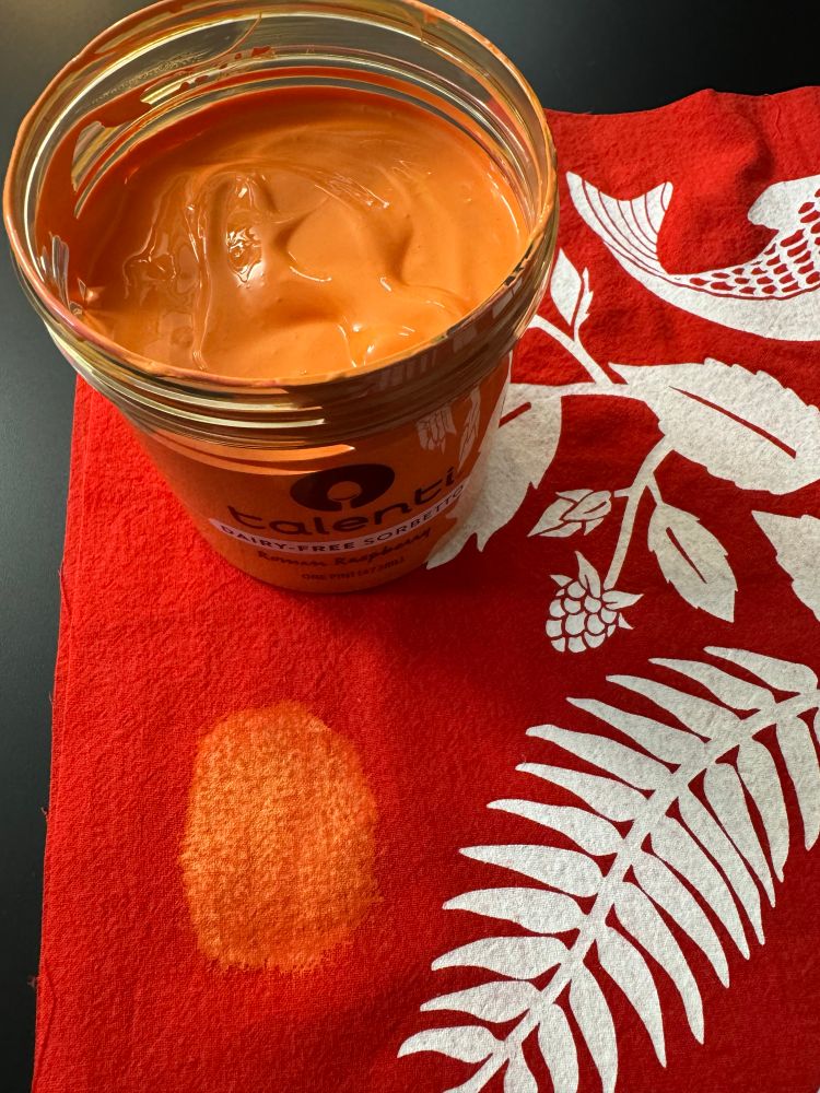 A photo from above of an open plastic sorbet pint container which is filled with peachy colored screen print ink. The container of ink sits on a piece of red fabric that has white printing on it and smudges of the peach colored ink sampled on it.