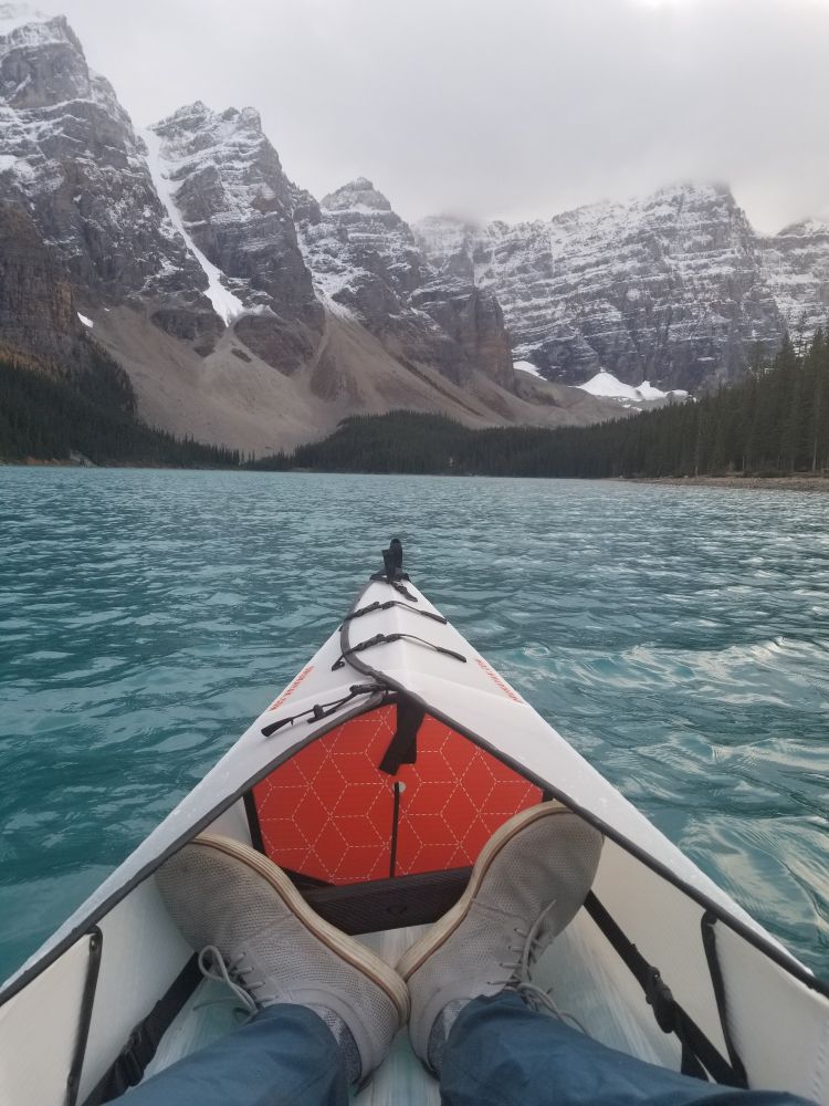 My view from my kayak as I floated around Morain Lake. My white and orange kayak with foggy mountains over the turquoise blue water.