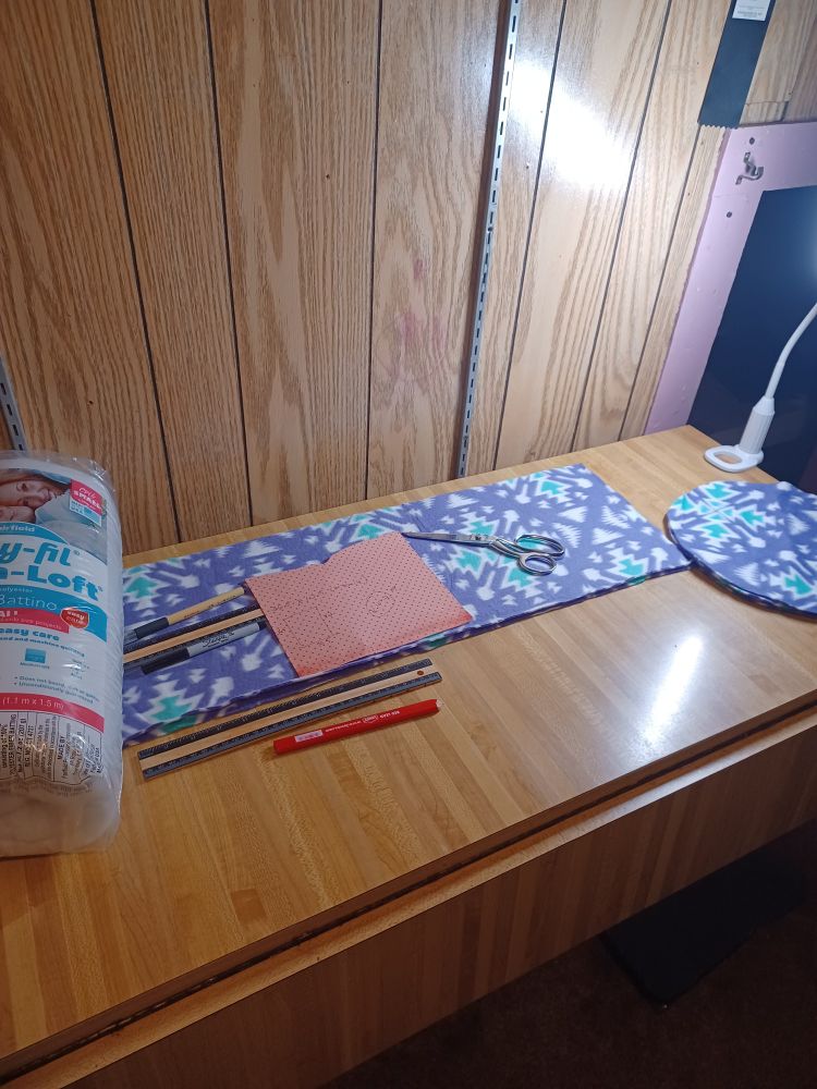 Photo of a sewing workspace with purple fleece cut into different shapes on it. Scissors, markers, a ruler and a bag of batting are set on top. 