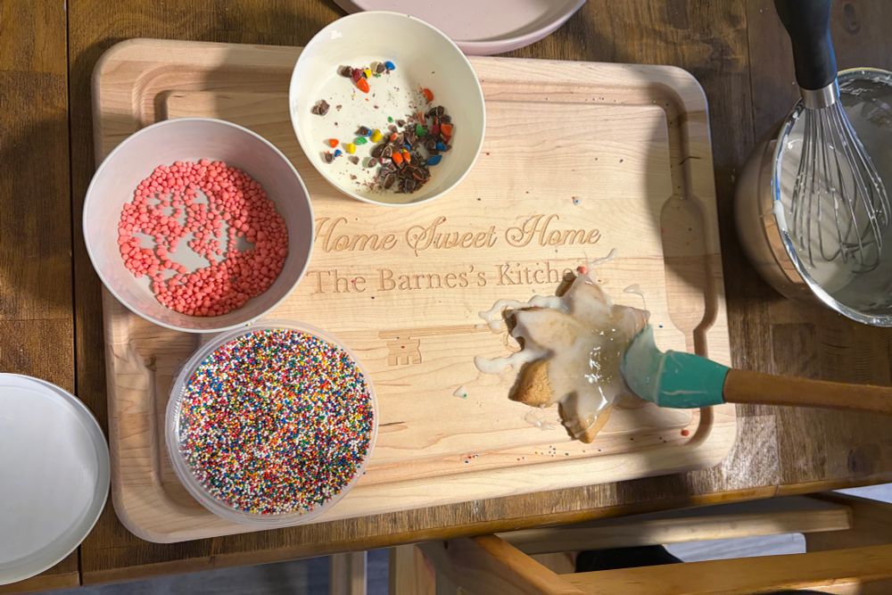 Sugar cookie decorations in a bunch of bowls on a cutting board that says “Home Sweet Home The Barnes’s Kitchen” — one cookie is being glazed.