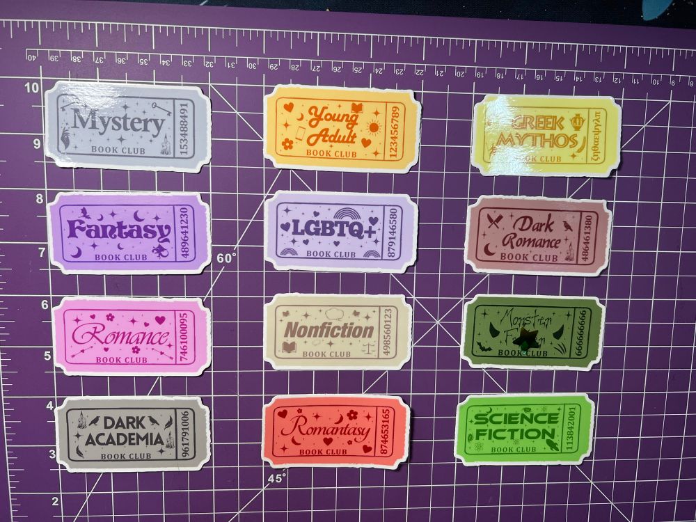 12 stickers on a purple background. Stickers are 3 inches by 1.5 inches and look like raffle tickets with the name of the genre, book club, and some doodles. 
Stickers include: mystery, young adult, Greek mythos, fantasy, lgbtq+, dark romance, romance, nonfiction, monster f**ker, dark academia, romantasy, and science fiction