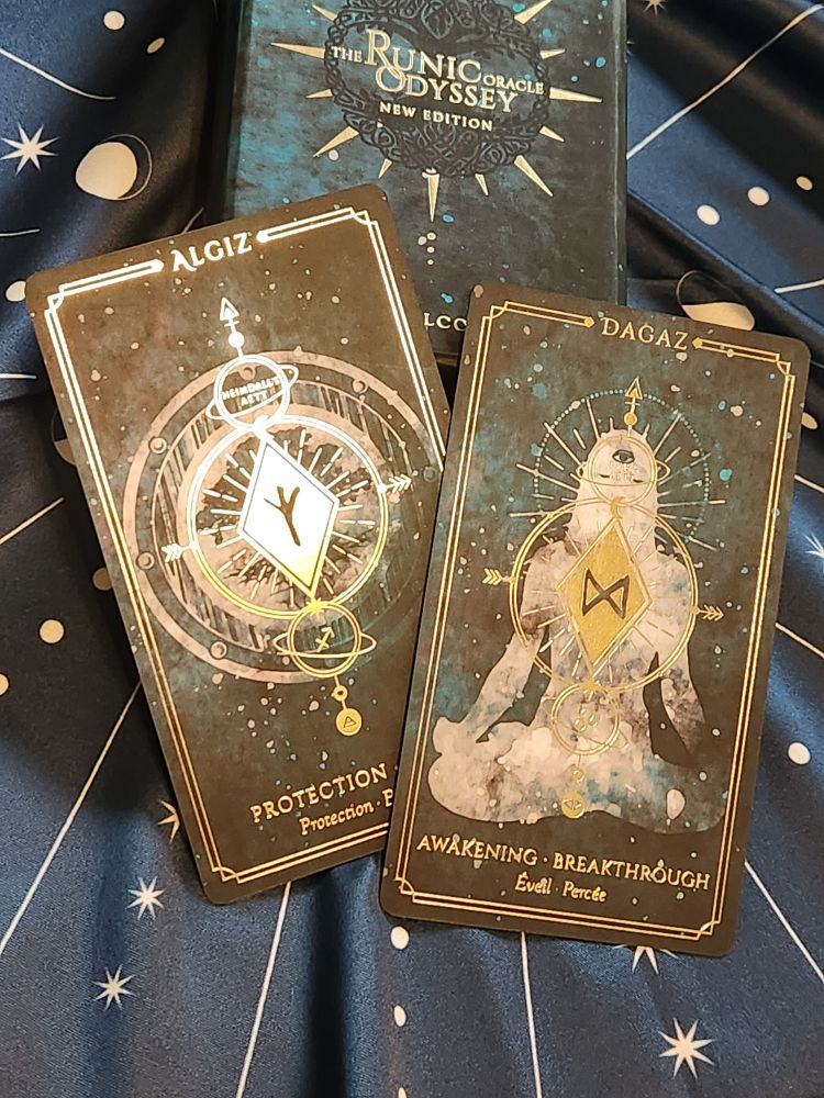two cards, both with gold details on dark green backgrounds. algiz, on the left, shows a round wooden shield. moon phases are around the edges. a large tree with wide canopy and visible roots is in the center. on top of the image is a gold diamond. the rune in the center has three branching lines at the top that connect to a single line. card text reads: "algiz. protection - shield." the word "shield" is covered by the other card, dagaz. this card has a silhouette of a human in the center. they sit cross legged with hands at their sides. no features are shown except for an opened third eye at the center of the forehead. the middle of the image has a gold diamond and rune like the other card. this rune is shaped like two triangles connected at one point. card text reads: "dagaz. awakening - breakthrough."
