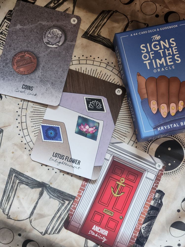 three cards in a diagonal line, with the deck box in the upper right. "coins" shows a cracked concrete background. two pieces of currency are scattered on it. one is a usa penny with the tails side up. the other is a usa dime with the heads side up. card text reads: "coins - good luck." to the right of this card is "lotus flower" card, showing a wooden surface that has several papers on top. one is a pale purple, the second is white, and the third is light brown. each has a different postage stamp of a flower, in various colors and styles. card text reads: "lotus flower - enlightenment." to the right of this card is the final one, "anchor". the image is focused on the closed door of a brick building. the door frame is white. the door itself is bright red. an anchor decoration hangs near the top. card text reads: "anchor - stability."