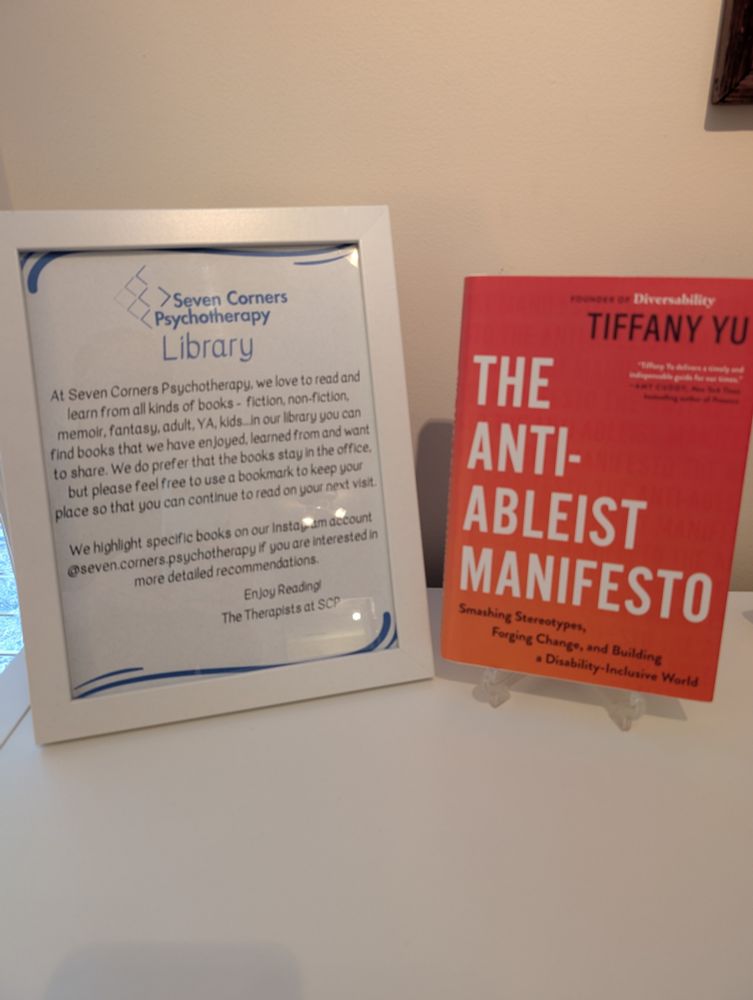 Photo of a sign that says "Seven Corners Psychotherapy Library" and explains that the therapists at the practice love to read and share the books they love but please leave them in the waiting room. The sign is next to a copy of "The Anti-Abelist Manifesto" by Tiffany Yu, the current highlighted book in honor of Disability Pride month 