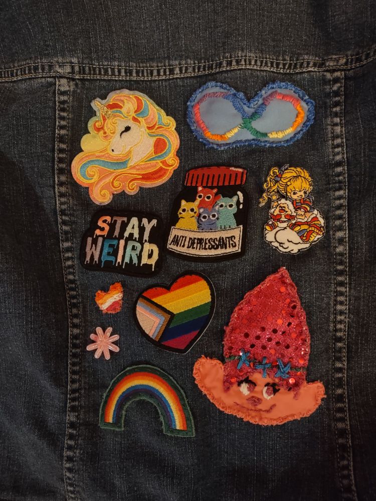 Photo of patches sewn to a jacket including a unicorn, the autism infinity symbol, a patch that reads 'stay weird', cats in a bottle labeled 'antidepressants', Rainbow Brite, lesbian pride heart, flower, progress flag heart, rainbow and Poppy from Trolls