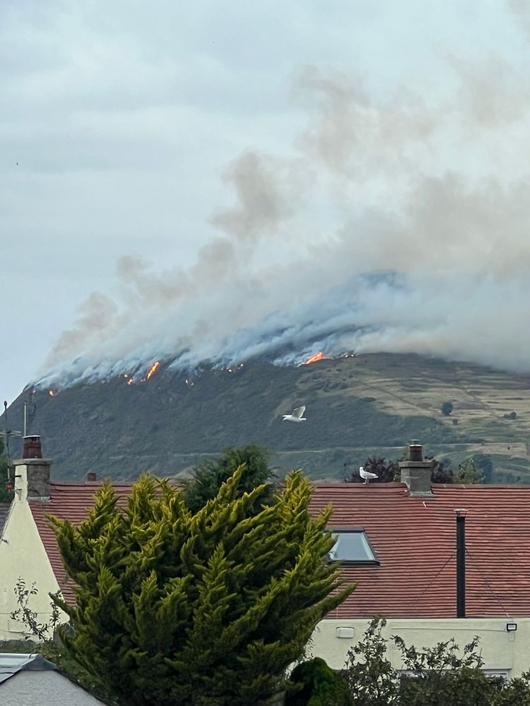A shot of Holyrood Park showing that the area around Arthur’s Seat is on fire; flames and clouds of billowing smoke are visible
