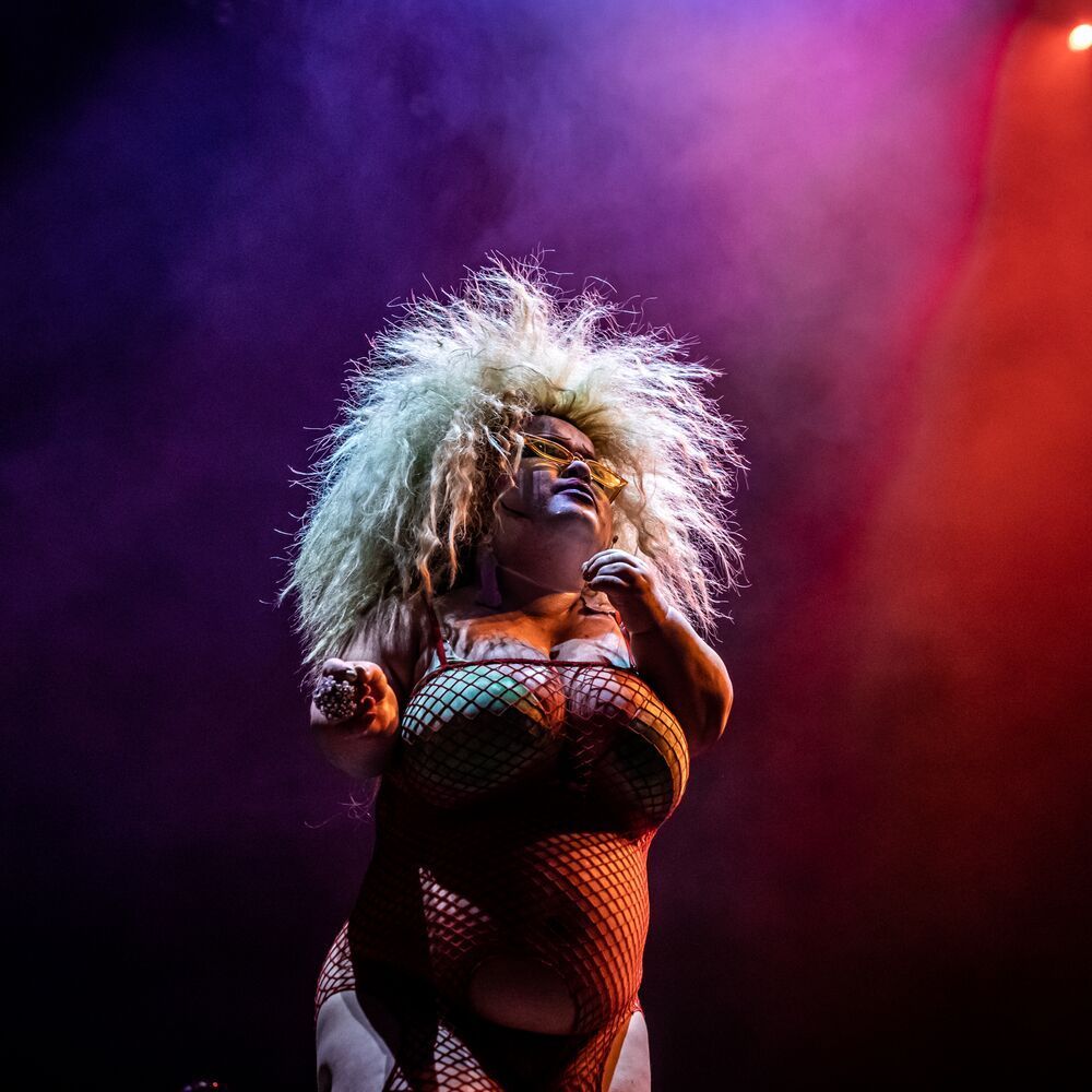 Midgitte Bardot stands in a fishnet dress and green bra with bright blonde hair that fans out against the purple and red lit backdrop.
