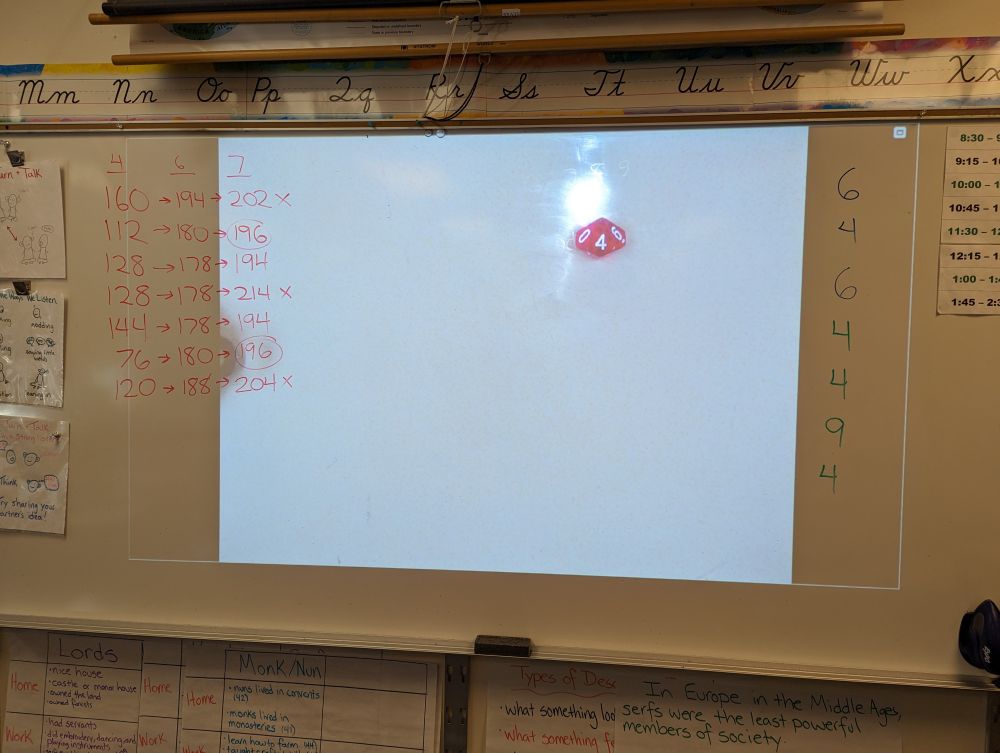 White board with a document camera projecting an image of a 10-sided dice showing the number 4. On one side of the white board, students' scores for a game are listed. On the other side is a record of all the dice rolls made during the game.