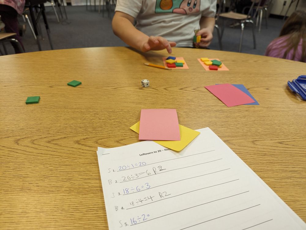 Table showing a math game in progress. On the far side of the table, a student is counting tiles that are being evenly grouped onto two small rectangles of construction paper. On the near side of the table is a recording sheet, a die, and additional small rectangles of construction paper.