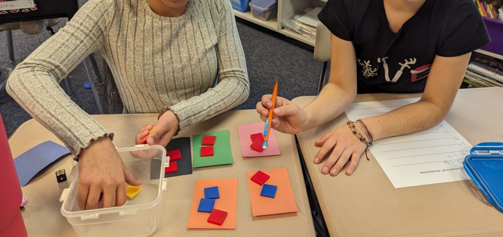 Two students playing a math game at their desk. They have square tiles and they are dividing them onto five small rectangles of construction paper.