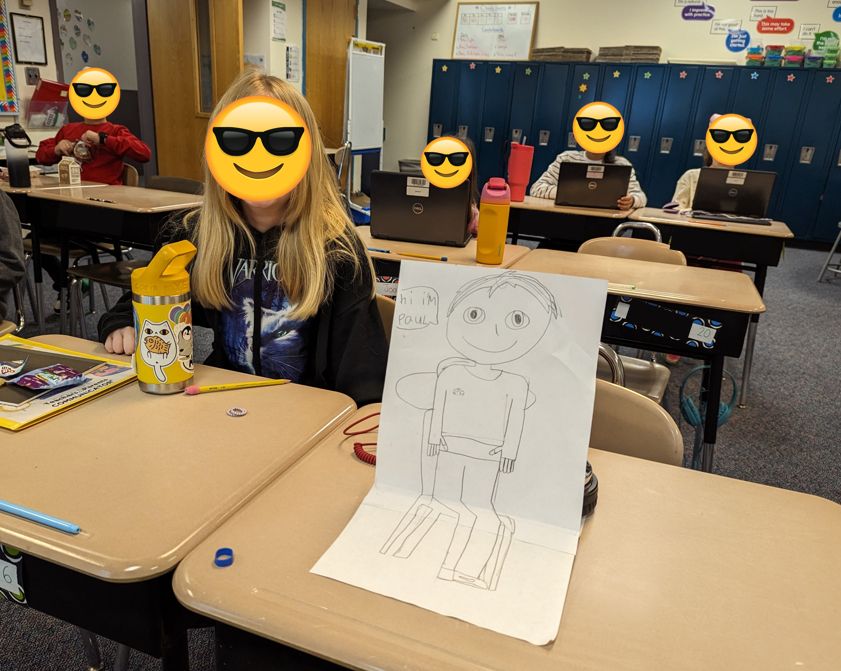 Photo of a classroom with desks and children. One desk has a piece of paper propped on top. The paper has a drawing of a child sitting in a chair with a speech bubble that says, "Hi, I'm Paul."