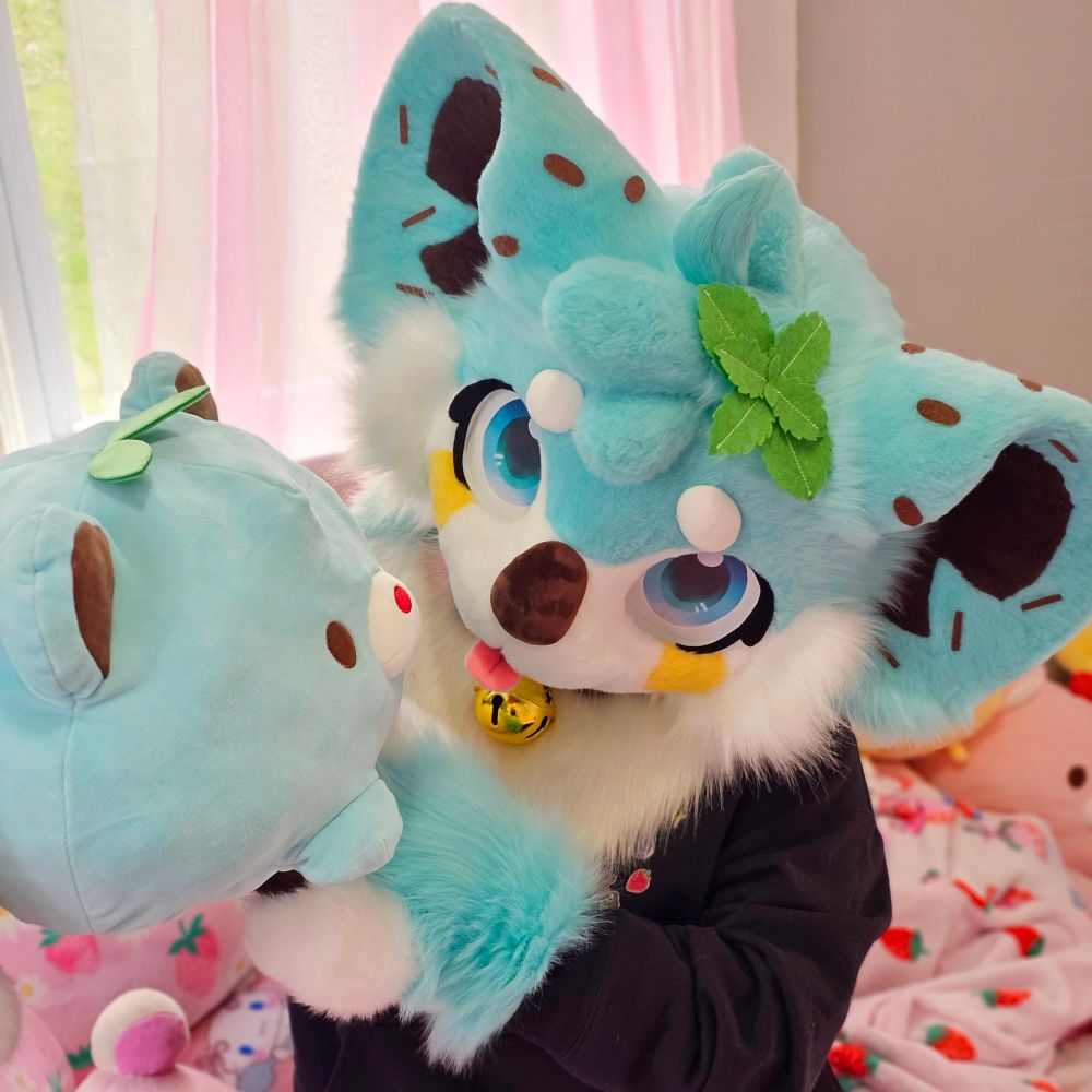 A mint chocolate chip koala fursuiter sitting on a pink bed holding a plushie to their side that unintentionally matches their fursuit. 