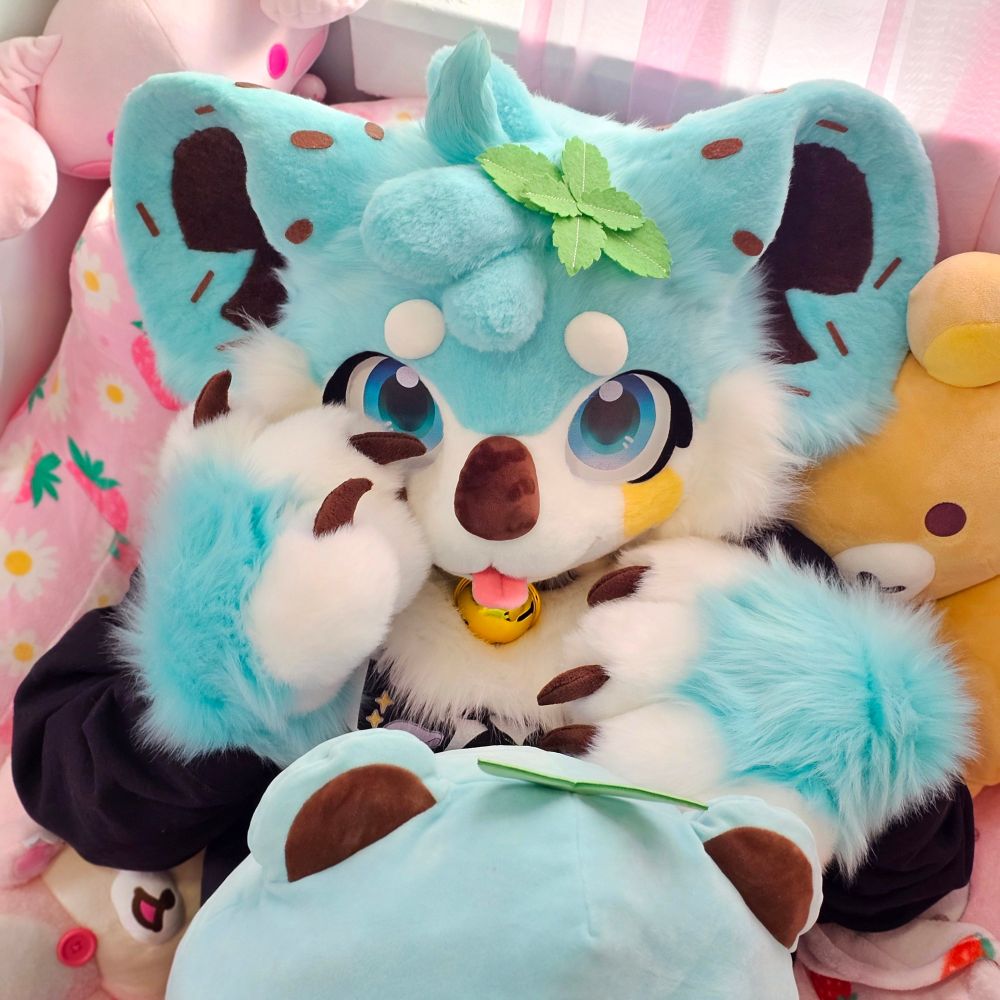 A mint chocolate chip koala fursuiter laying on a pink bed with plushies surrounding them. One of their paws is rested on their cheek, and the other on their chest. 