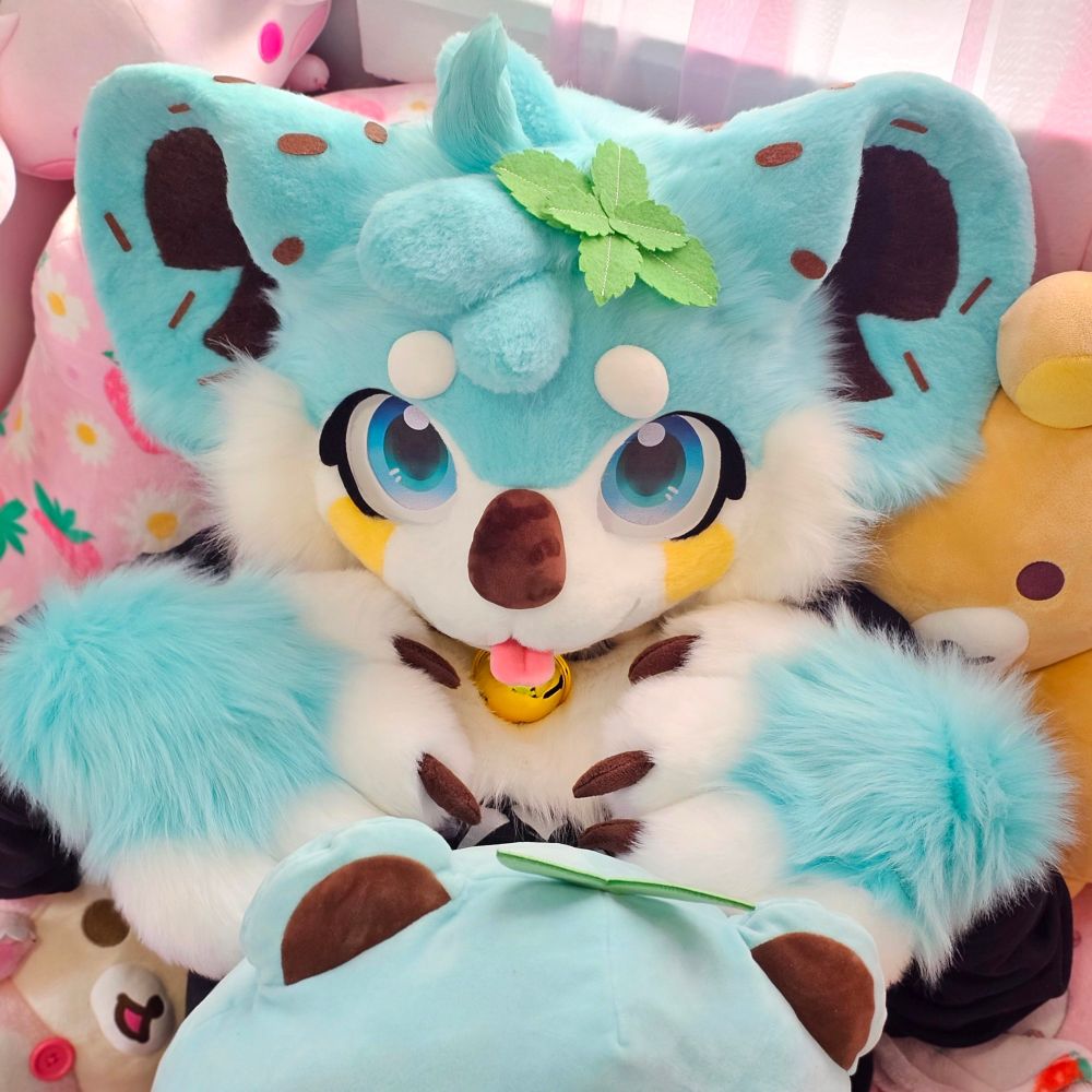 A mint chocolate chip koala fursuiter laying on a pink bed with plushies surrounding them. Both of their paws are rested on their chest. 